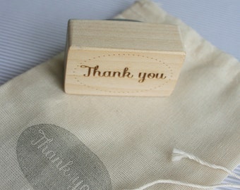 Puede incluir: Un sello de madera con las palabras "Gracias" grabadas. El sello está sobre una bolsa de tela blanca con cordón.