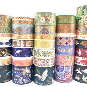 Può includere: Una collezione di venticinque rotoli di nastro adesivo decorativo washi impilati in cinque colonne. I nastri presentano vari motivi tra cui gru, ventagli, disegni floreali e geometrici nei colori oro, blu, verde, rosa e nero.