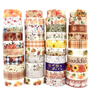 May include: Four vertical stacks of decorative washi tape rolls featuring autumn-themed patterns including pumpkins, sunflowers, mushrooms, fall leaves, plaid prints, and the word thankful printed on one of the rolls in a brown script font.