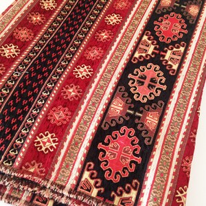 Puede incluir: Tela estampada en rojo, negro y dorado con un dise&ntilde;o geom&eacute;trico repetitivo. La tela tiene una textura tejida y un borde con flecos.