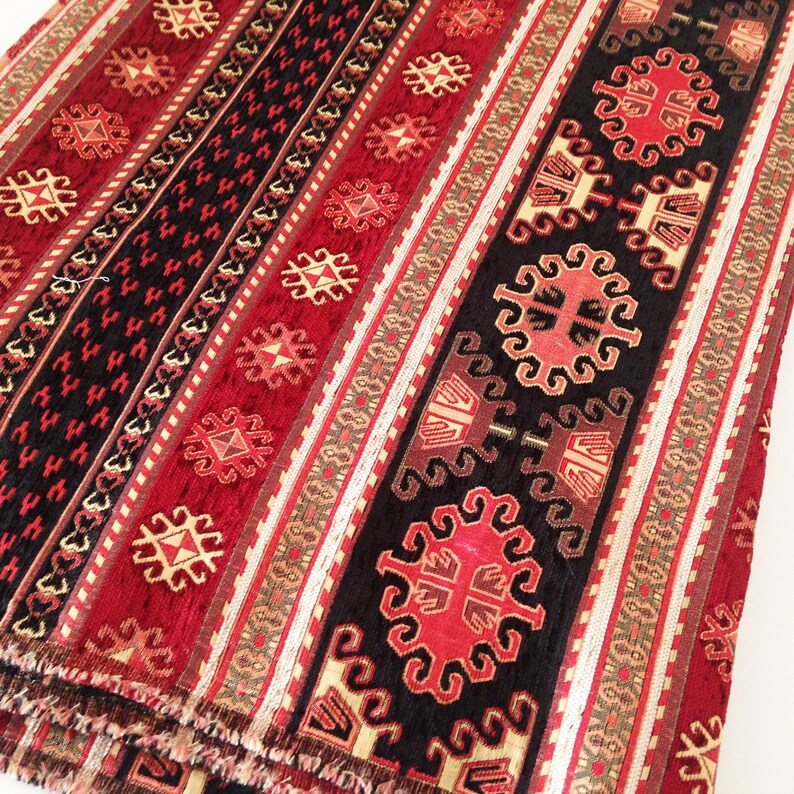 Puede incluir: Tela estampada en rojo, negro y dorado con un dise&ntilde;o geom&eacute;trico repetitivo. La tela tiene una textura tejida y un borde con flecos.