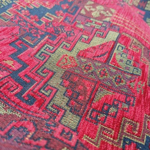 May include: Close-up of a red fabric with a gold and blue patterned design. The fabric has a textured surface and a repeating geometric pattern.