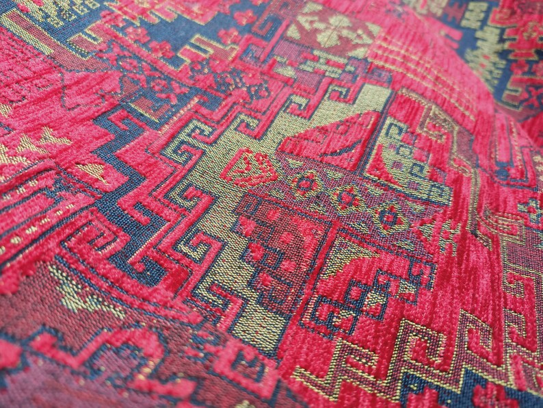 May include: Close-up of a red fabric with a gold and blue patterned design. The fabric has a textured surface and a repeating geometric pattern.