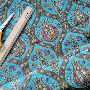 May include: A bright turquoise fabric featuring a repeating floral pattern in shades of dark brown, red, and cream. A pair of orange-handled scissors and a wooden ruler measuring 30 centimeters are placed on the fabric to show scale.