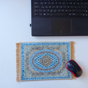 Op de afbeelding: Een rechthoekige muismat met een complex oosters tapijtpatroon in blauw en goud, voorzien van een centraal medaillon en een decoratieve rand. De muismat heeft goudkleurige franjes aan de linker- en rechterkant en ligt naast een zwarte computermuis.