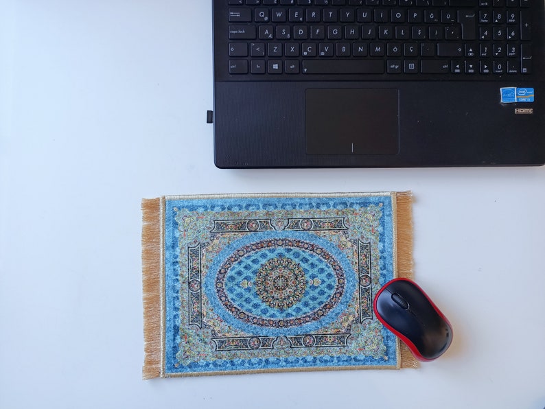 Op de afbeelding: Een rechthoekige muismat met een complex oosters tapijtpatroon in blauw en goud, voorzien van een centraal medaillon en een decoratieve rand. De muismat heeft goudkleurige franjes aan de linker- en rechterkant en ligt naast een zwarte computermuis.
