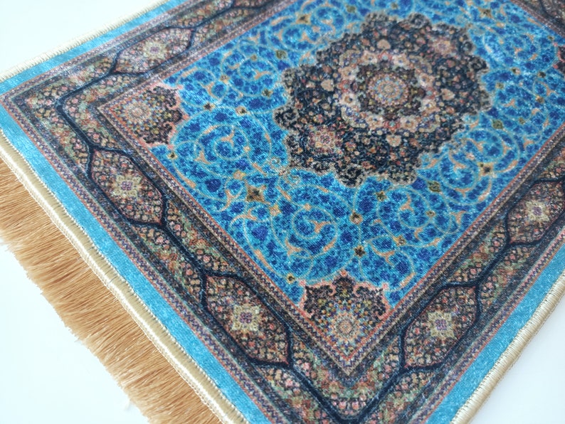 K&ouml;nnte beinhalten: Ein rechteckiger Teppich mit einem leuchtend blauen Hintergrund und komplizierten schwarzen und goldenen Blumenmustern. Der Teppich hat ein zentrales Medaillon-Design, einen detaillierten Rand und lange, hellbraune Fransen an den kurzen Kanten.