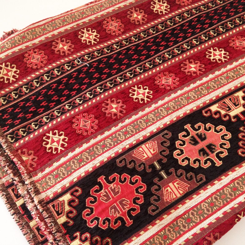 Puede incluir: Tela estampada en rojo, negro y dorado con un dise&ntilde;o geom&eacute;trico. La tela tiene una textura tejida y un borde con flecos.