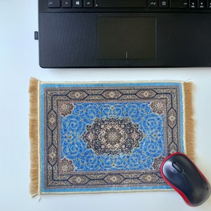K&ouml;nnte beinhalten: Ein rechteckiges Mauspad mit einem komplexen blauen, schwarzen und beigen Blumenmuster und einem zentralen Medaillon-Design. Das Mauspad hat hellbraune Fransen an den linken und rechten R&auml;ndern und liegt unter einem schwarzen Laptop.