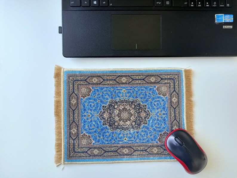 K&ouml;nnte beinhalten: Ein rechteckiges Mauspad mit einem komplexen blauen, schwarzen und beigen Blumenmuster und einem zentralen Medaillon-Design. Das Mauspad hat hellbraune Fransen an den linken und rechten R&auml;ndern und liegt unter einem schwarzen Laptop.