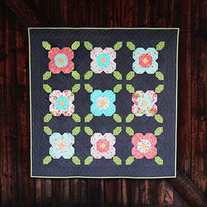 May include: A quilt with a dark gray background and nine colorful flower-shaped patches. The flowers are made of pink, blue, yellow, and white fabric. The quilt is hanging on a wooden wall.