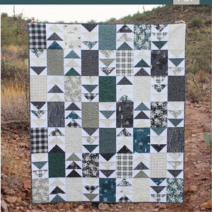 Op de afbeelding: Een quiltpatroon genaamd "Take Me Home" #130, met een patchwork-ontwerp van grijze, witte, zwarte en teal stof. De quilt is gemaakt van vierkanten en driehoeken, met een verscheidenheid aan patronen en texturen. De quilt wordt getoond tegen een achtergrond van rotsen en woestijnplanten.