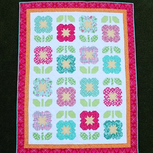 Op de afbeelding: Een wit quilt met een bloemenpatroon. Het quilt is gemaakt van vierkanten met verschillende bloemenpatronen in tinten roze, blauw, groen en geel. Het quilt is afgewerkt met een roze, oranje en witte bies.