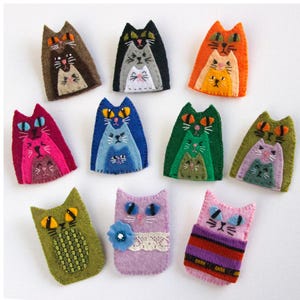 May include: A collection of eleven felt cat-shaped ornaments in various colours including brown, black, orange, pink, blue, green, olive, and purple. Each ornament features embroidered facial details and layered cat designs, with some featuring lace or fabric.
