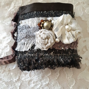 Puede incluir: Un brazalete de muñeca de tela marrón y blanco con un diseño floral decorativo y lazos de cinta marrón. El brazalete está hecho de varias telas y texturas, incluyendo tweed, encaje y arpillera. La flor está adornada con perlas y cuentas.