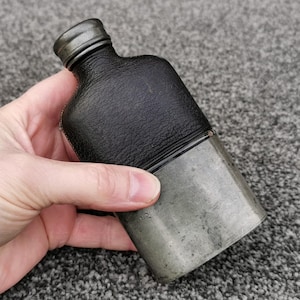 Könnte beinhalten: Eine Vintage-Flasche mit einem schwarzen, lederbezogenen Oberteil und einem silberfarbenen Metallsockel. Die Flasche hat einen kleinen, runden, silberfarbenen Deckel. Das Leder hat ein strukturiertes Aussehen. Die Flasche wird in einer Hand gehalten.