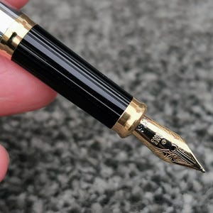 May include: Close-up of a fountain pen with a gold nib and accents. The pen has a black, ridged barrel and a silver cap. The nib is engraved with decorative patterns and the word "BOSS".