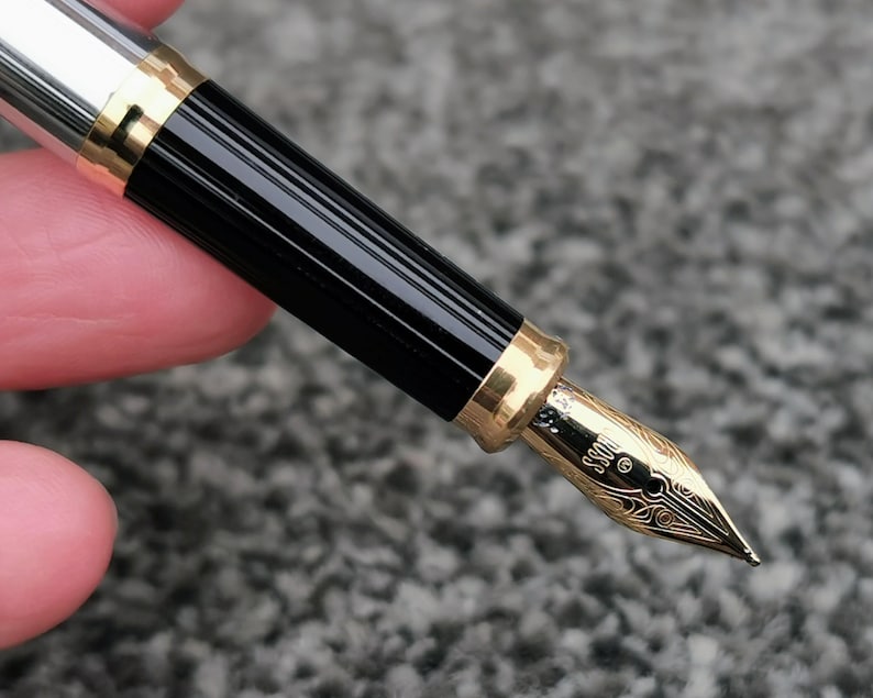 May include: Close-up of a fountain pen with a gold nib and accents. The pen has a black, ridged barrel and a silver cap. The nib is engraved with decorative patterns and the word "BOSS".