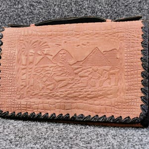 May include: A light brown leather clutch purse with a black trim. The purse has an embossed design of pyramids and palm trees.