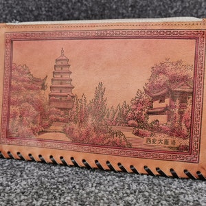May include: A brown leather zipped pouch with a stitched border. The pouch features an embossed image of a pagoda and other buildings with the text "西安大雁塔" in Chinese.