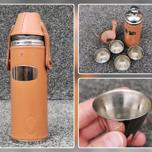 May include: A cylindrical stainless steel thermos flask wrapped in tan leather with a matching leather cap and strap. The set includes four small stainless steel cups that nest together, featuring a debossed emblem on the leather exterior.