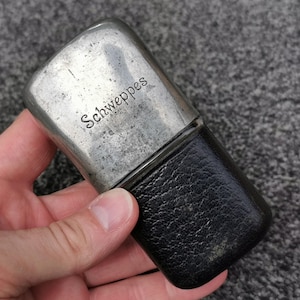 May include: A rectangular metal flask with a weathered silver-tone finish and the word Schweppes engraved on the front. The lower portion of the flask is covered in a textured black material, creating a two-tone appearance with rounded edges.