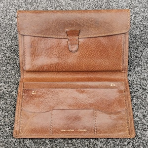 May include: Open brown leather wallet with a flap and buckle closure. The wallet is open, showing multiple card slots and a larger compartment. The words "REAL LEATHER" are visible. The wallet is a warm brown colour.