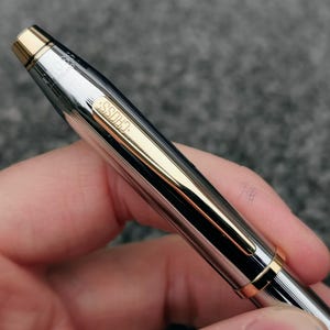 May include: A silver and gold Cross pen with a ribbed design. The pen has gold accents on the tip, clip, and band. The word "CROSS" is engraved on the pen's side. The pen is held in a hand against a gray background.