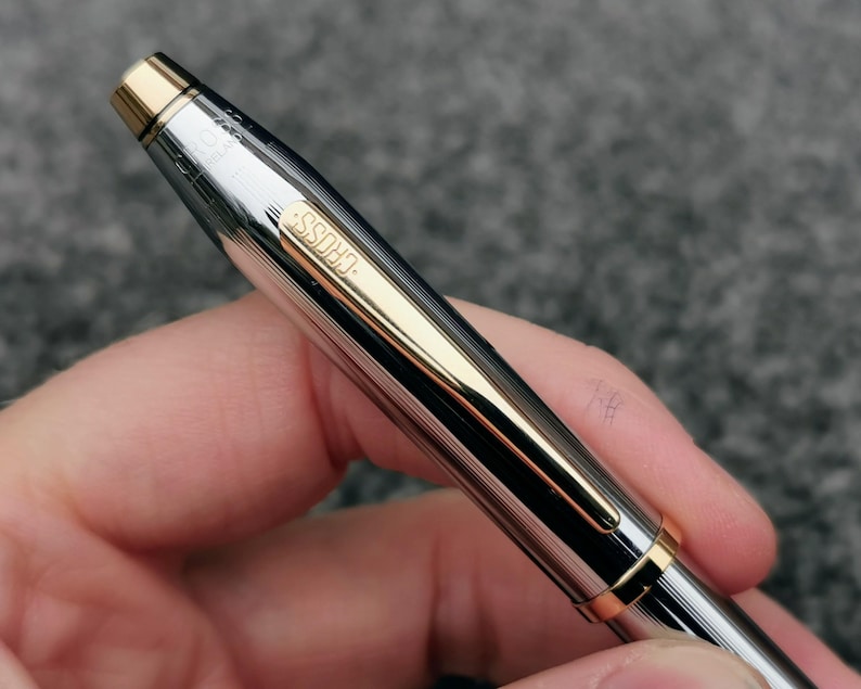 May include: A silver and gold Cross pen with a ribbed design. The pen has gold accents on the tip, clip, and band. The word "CROSS" is engraved on the pen's side. The pen is held in a hand against a gray background.