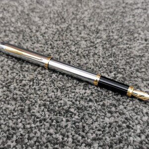 May include: A silver fountain pen with gold accents and a black grip. The pen has a gold nib and a clip. The pen is lying on a gray, textured surface.