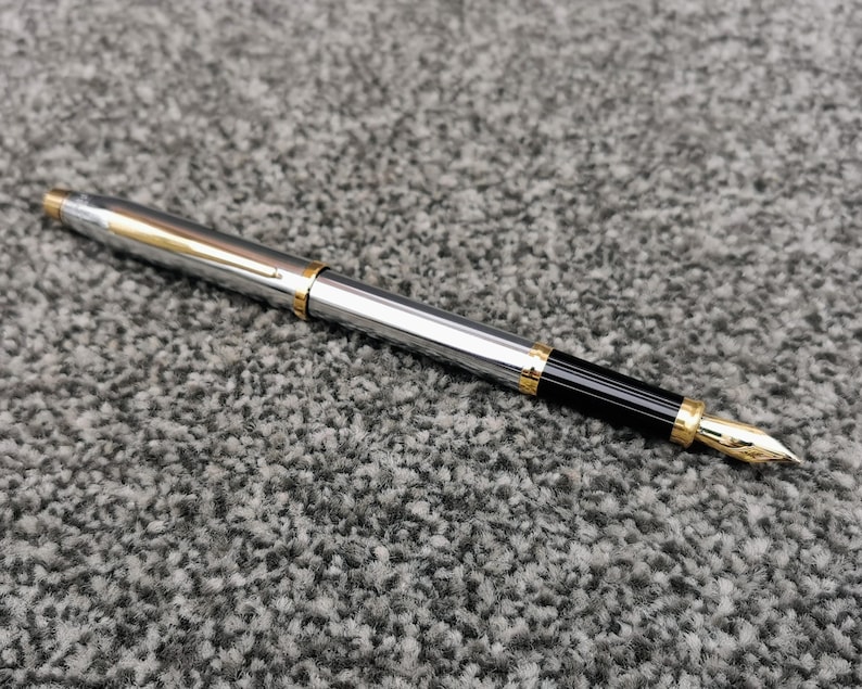 May include: A silver fountain pen with gold accents and a black grip. The pen has a gold nib and a clip. The pen is lying on a gray, textured surface.