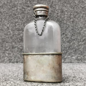 Könnte beinhalten: Ein Flachmann aus klarem Glas mit einem silberfarbenen Metallsockel und einem passenden Schraubverschluss. Eine dünne silberne Kette verbindet den Verschluss mit dem Flaschenhals, der eine abgerundete Oberseite und einen rechteckigen Körper hat.
