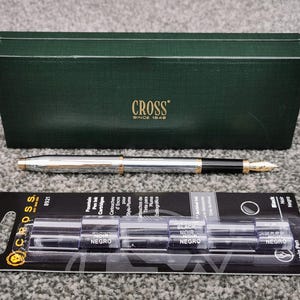 May include: A silver and gold fountain pen with a black grip and gold nib, next to a green box with the word "CROSS" and a package of black ink cartridges. The pen and box are on a gray surface.