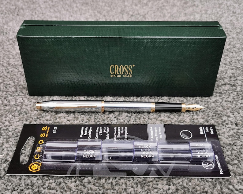 May include: A silver and gold fountain pen with a black grip and gold nib, next to a green box with the word "CROSS" and a package of black ink cartridges. The pen and box are on a gray surface.