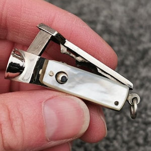 Può includere: Un piccolo tagliaunghie argentato con intarsio in madreperla. Il tagliaunghie ha un design a cerniera con una leva e un piccolo anello per il fissaggio. La madreperla ha una lucentezza perlacea.