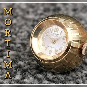 May include: A white ceramic mug with a black handle and the text "But First, Coffee" in a black, handwritten-style font. The mug is on a white surface.