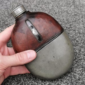 Puede incluir: Una cantimplora metálica redondeada con una funda de cuero marrón oscuro en la parte superior y una base de metal gris lisa y sin pulir. La cantimplora incluye una pequeña ventana vertical en el cuero y un tapón metálico plateado de rosca.