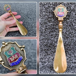 May include: A brass-colored shoehorn with a decorative handle featuring a shield emblem. The shield has a red, green, and white design with the word "WINDSOR" in blue. The shoehorn is approximately 10 inches long.