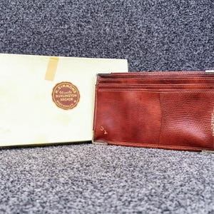 May include: A reddish-brown leather wallet with a textured surface, rectangular shape and metal corner accents. Beside it is a cream box with a circular logo reading "H. Simmons 60 & 61 Burlington Arcade Piccadilly."