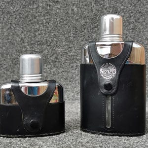 May include: Two silver-toned metal flasks with polished finishes, each encased in a black leather sleeve with a snap-button closure. The larger flask features a vertical cutout revealing the metal surface and a circular logo with the text SAG.