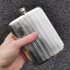 May include: A silver hip flask with a screw-on cap. The flask has a rectangular shape with vertical striped detailing. The flask is held in a hand against a grey background.