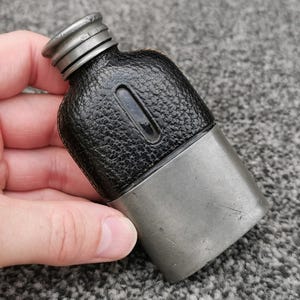 Könnte beinhalten: Ein kleiner Flachmann mit einem silberfarbenen Metallsockel und einem oberen Teil aus schwarzem, strukturiertem Lederimitat. Der Flachmann hat einen schmalen Hals mit Schraubverschluss und ein vertikales Sichtfenster auf der Vorderseite.