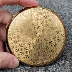 May include: A round gold-tone metal compact mirror featuring an intricate, repeating geometric floral pattern etched across the entire surface. The compact has a polished finish with a metallic sheen and a small side latch for opening.