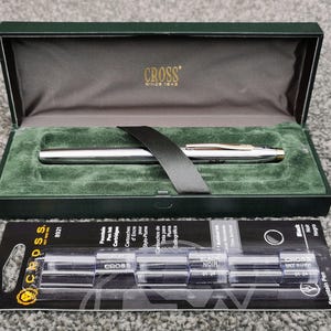 May include: A silver fountain pen with gold accents rests in a green velvet-lined box. The box is open, revealing the pen and a black ribbon. Below the box is a package of black ink cartridges. The box has the word "CROSS" printed on it.