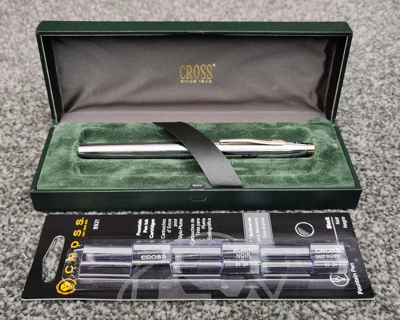 May include: A silver fountain pen with gold accents rests in a green velvet-lined box. The box is open, revealing the pen and a black ribbon. Below the box is a package of black ink cartridges. The box has the word "CROSS" printed on it.