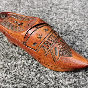 Op de afbeelding: Een houten schoenvormige doos met ingewikkelde gravures. De doos heeft een warme bruine kleur en is voorzien van het woord "ANVER". Decoratieve patronen en sterachtige ontwerpen zijn ook in het hout gesneden.