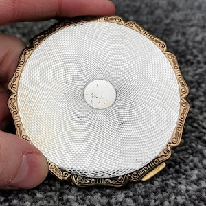 May include: A vintage, round compact with a silver-toned, textured surface and a gold-colored, ornate border. The compact has a small, circular indentation in the center. The compact is held by a hand.