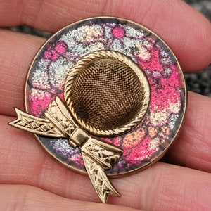 May include: A gold-tone brooch with a pink and white patterned hat design. The hat has a mesh crown and a gold bow.