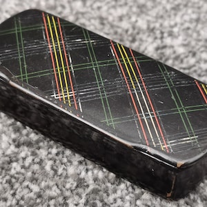 Peut inclure: Une boîte rectangulaire noire avec un motif écossais en vert, jaune, rouge et blanc. La boîte a un dessus et un dessous légèrement arrondis.