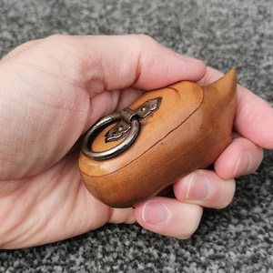 May include: A small, wooden box with a rounded shape and a pointed end. The box is light brown with a metal ring and clasp on top. The box is held in a hand.